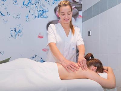 Kobieta otrzymująca masaż pleców od masażystki w jasnym pokoju zabiegowym z kwiatową ścianą.