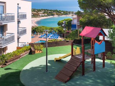 Plac zabaw dla dzieci ze zjeżdżalnią obok budynków hotelowych, z widokiem na plażę i morze w tle