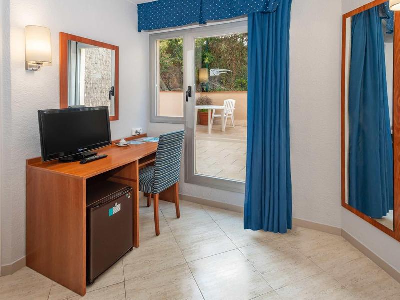 Pokój hotelowy z biurkiem, krzesłem, telewizorem, małą lodówką i dostępem do tarasu z stołem i krzesłami.