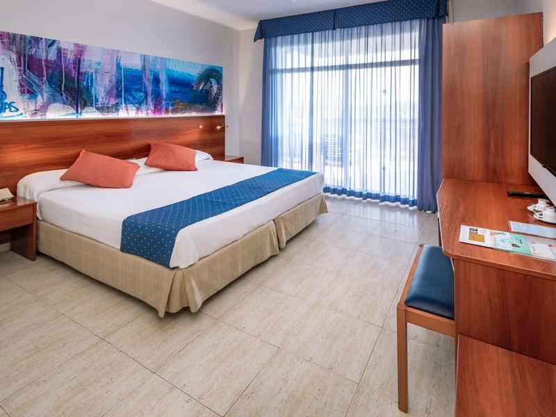 Jasny pokój hotelowy z dużym łóżkiem, biurkiem i oknem sięgającym podłogi z zasłonami.