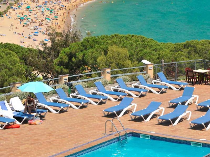 Basen hotelowy z leżakami z widokiem na zatłoczoną piaszczystą plażę i niebieskie morze.