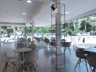 Café moderne et lumineux avec des chaises blanches et de grandes fenêtres donnant sur des arbres et des parkings.