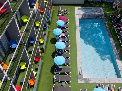 Vue de dessus d'une piscine rectangulaire avec chaises longues et parasols colorés à côté d'un bâtiment hôtelier.