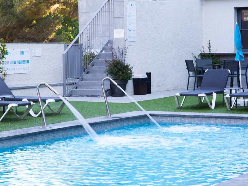Piscine extérieure rafraîchissante avec jets d'eau et chaises longues près d'un bâtiment hôtelier moderne.