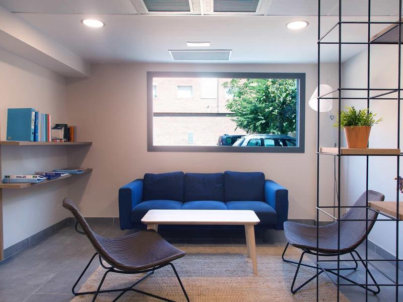 Espace d'attente moderne avec canapé bleu, deux fauteuils noirs et une table blanche sous une fenêtre.