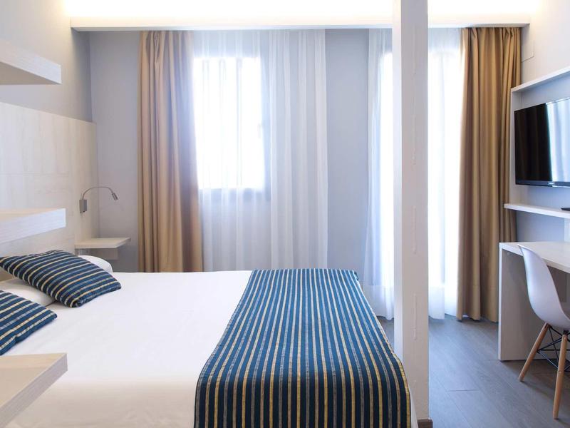 Chambre d'hôtel moderne avec lit double, couvre-lit à rayures et grande fenêtre avec rideaux.