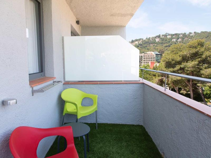 Petit balcon avec pelouse artificielle, deux chaises colorées et vue sur des collines verdoyantes.