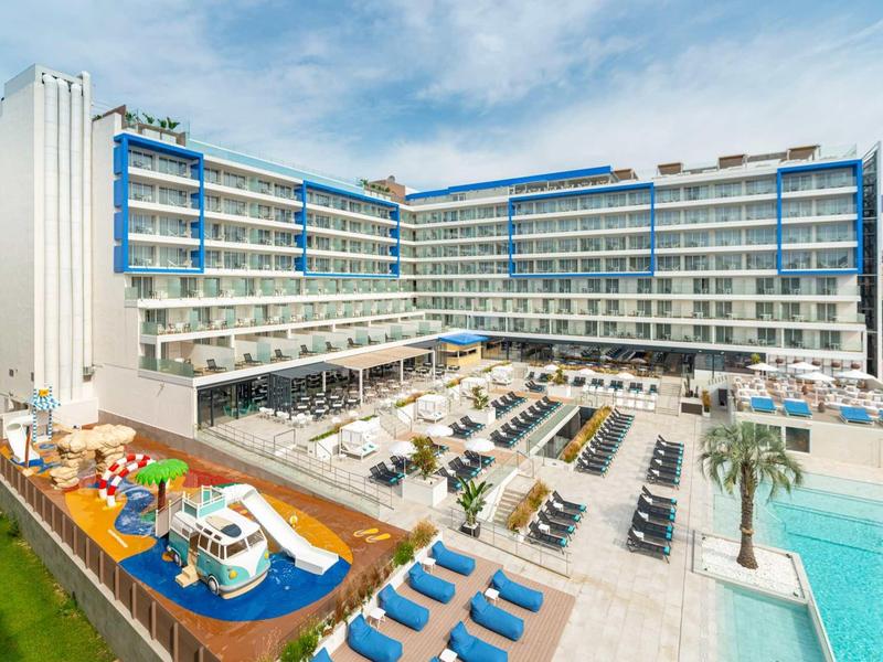 Hotel moderno con piscina, terrazza soleggiata e area giochi per bambini sotto un cielo limpido.