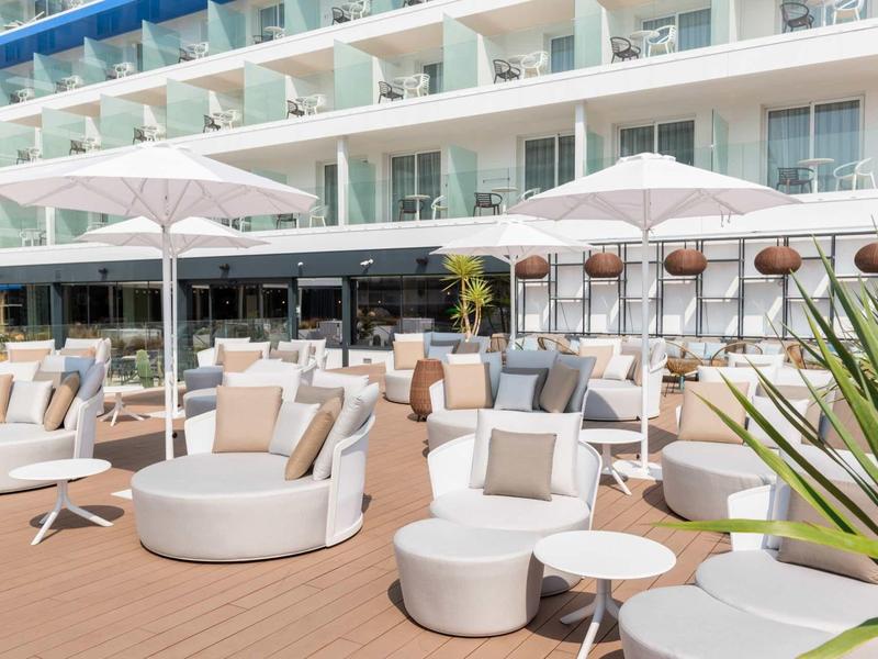 Lounge esterna moderna con poltrone bianche, tavoli e ombrelloni davanti alla terrazza dell'hotel.