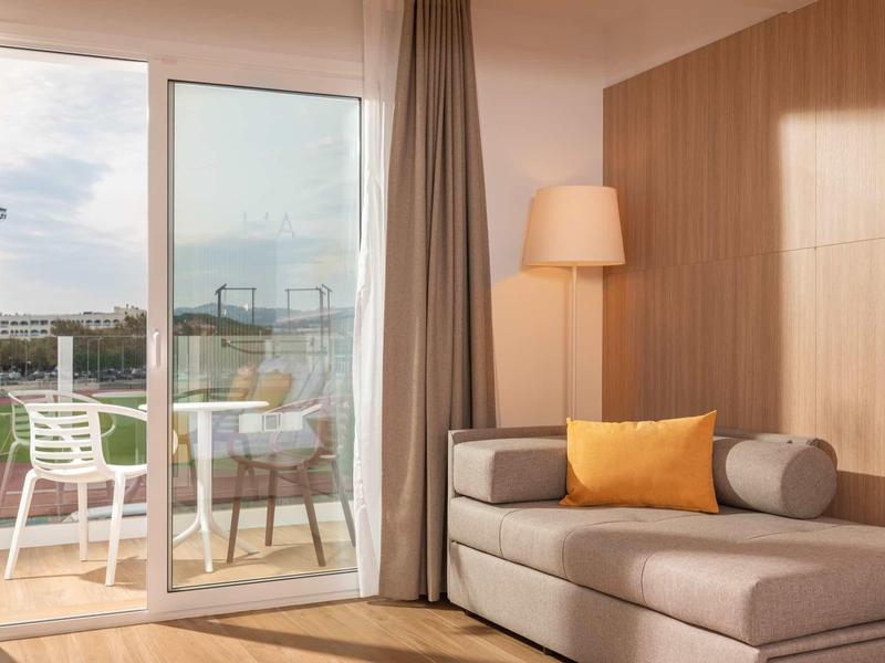 Camera d'hotel luminosa con divano, cuscini e vista sul balcone con sedie.