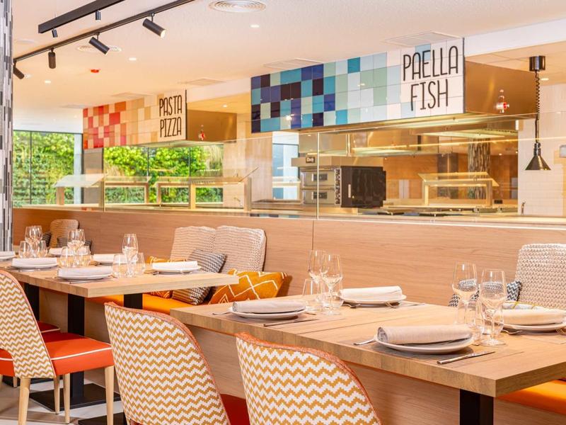 Ristorante luminoso con tavoli apparecchiati e cucina a vista con cartello 'Paella Fish'.