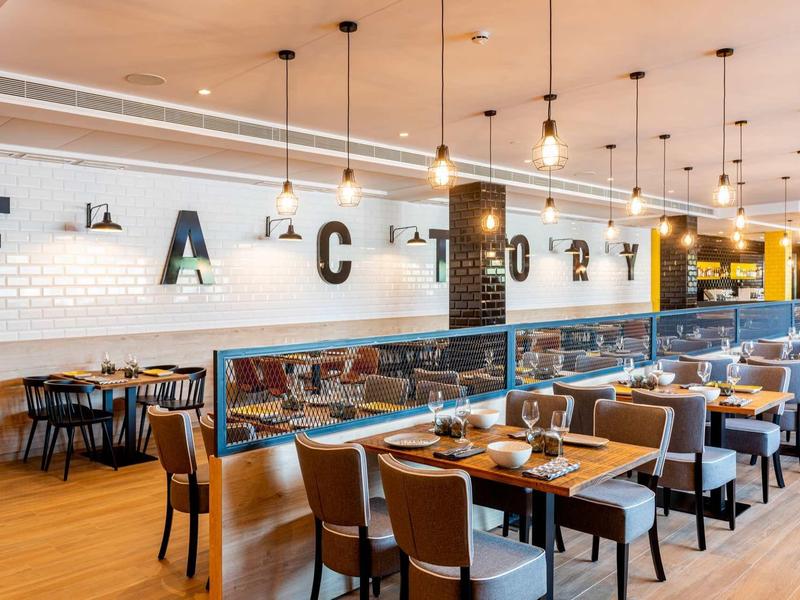 Ristorante moderno con pavimento in legno, sedie marroni e pareti bianche con la scritta FACTORY.