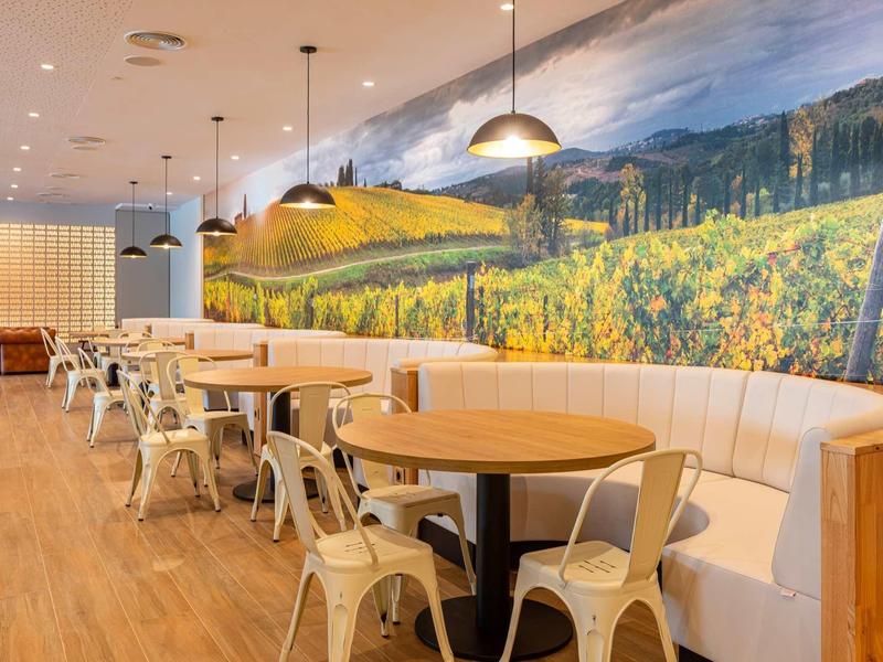 Sala da pranzo moderna con pavimento in legno, tavoli rotondi, sedie bianche e grande murale paesaggistica.