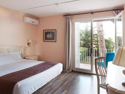 Světly hotelový pokoj s manželskou postelí, psacím stolem, židlí a balkonem s výhledem na zeleň.