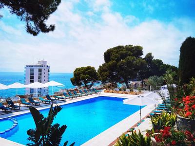 Venkovní bazén vedle květin a rostlin s výhledem na moře a budovy pod čistou oblohou.