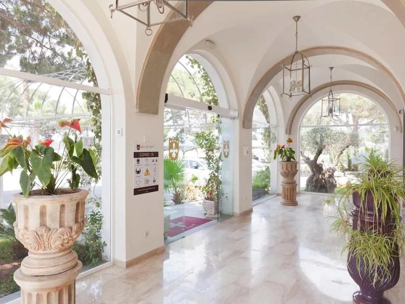 Světlá hotelová chodba s mramorovou podlahou, velkými okny a dekorativními rostlinami ve vázách.