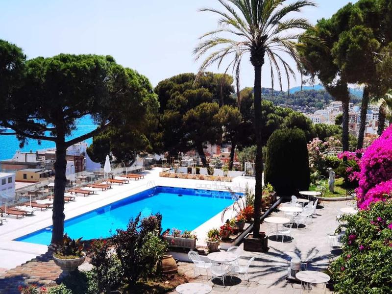 Pohled na hotelový bazén s palmami, lehátky a ženou na balkoně.