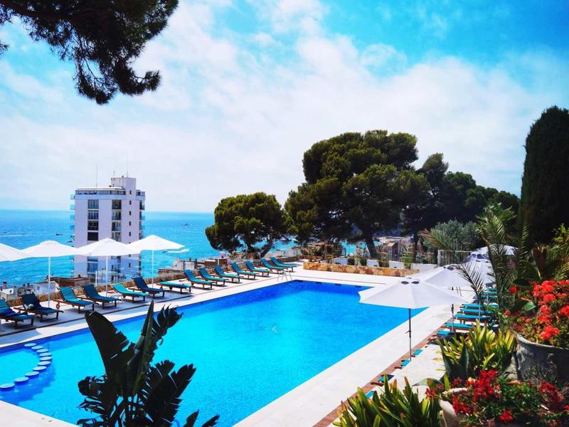 Venkovní bazén vedle květin a rostlin s výhledem na moře a budovy pod čistou oblohou.