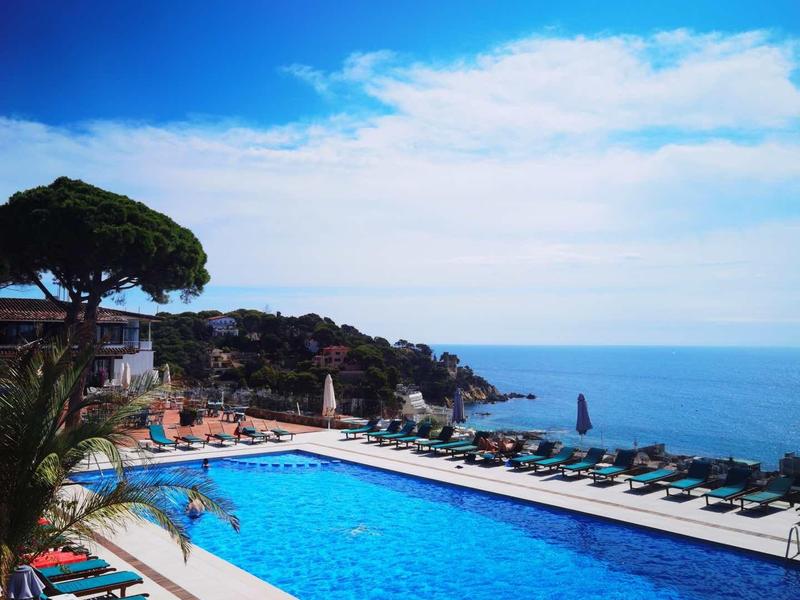 Velký venkovní bazén s lehátky, palmami a výhledem na moře pod modrou oblohou.