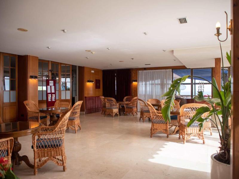 Světlá hotelová lobby s ratanovým nábytkem, velkými okny a rostlinami.