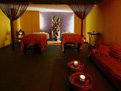 Warmly lit spa room with two massage tables, candles, and a seating area with cushions.