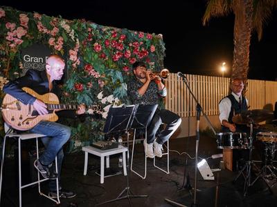 Tre musicisti suonano su un palco serale con una parete di fiori sullo sfondo.