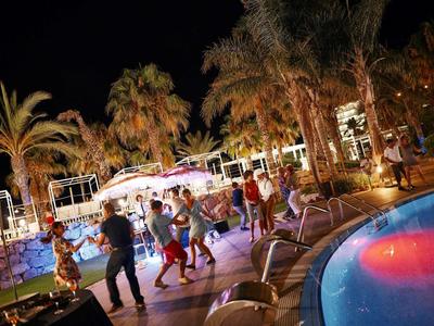 Persone che ballano di notte sotto palme accanto a una piscina illuminata in un'atmosfera rilassante del resort.
