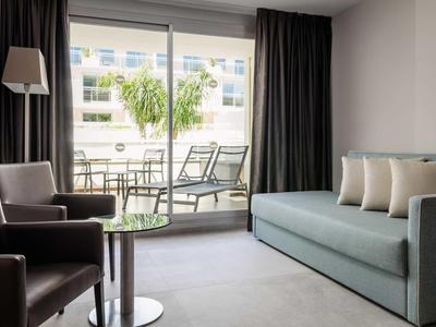 Modern hotel room with sofa, armchair, glass table, and balcony with chairs and palm view.