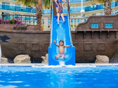 Children enjoying a blue water slide landing into a pool at a pirate ship-themed water park.