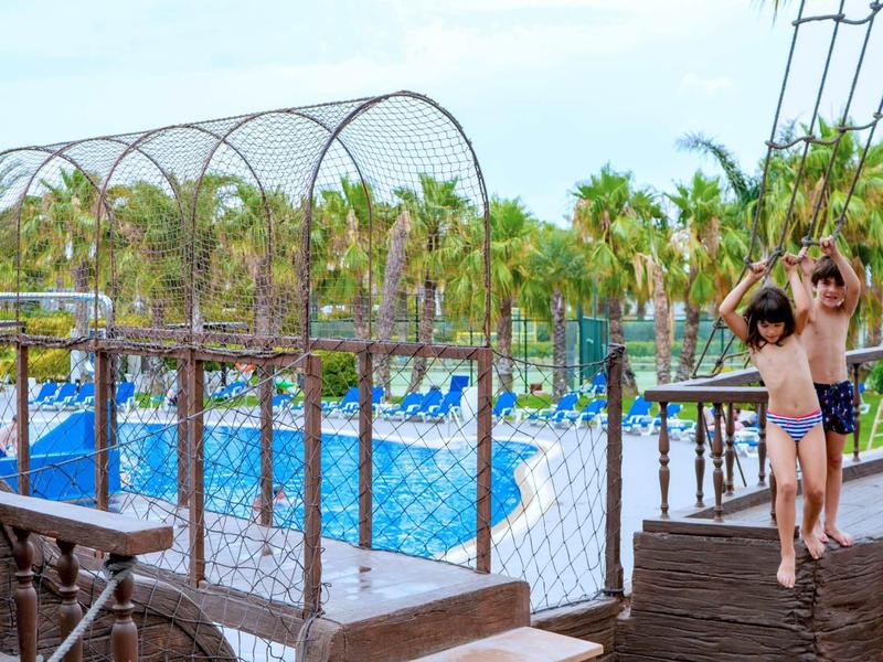 Kids walking on a wooden bridge near a fenced pool surrounded by palm trees and loungers.