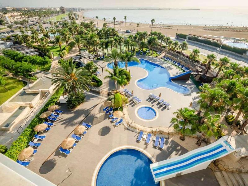 Aerial view of a hotel pool area with palm trees, lounge chairs, and a water slide near the beach.