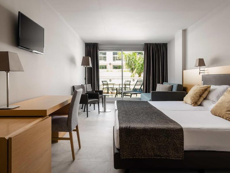 Modern hotel room with double bed, desk, TV, and patio door to balcony with outdoor furniture.