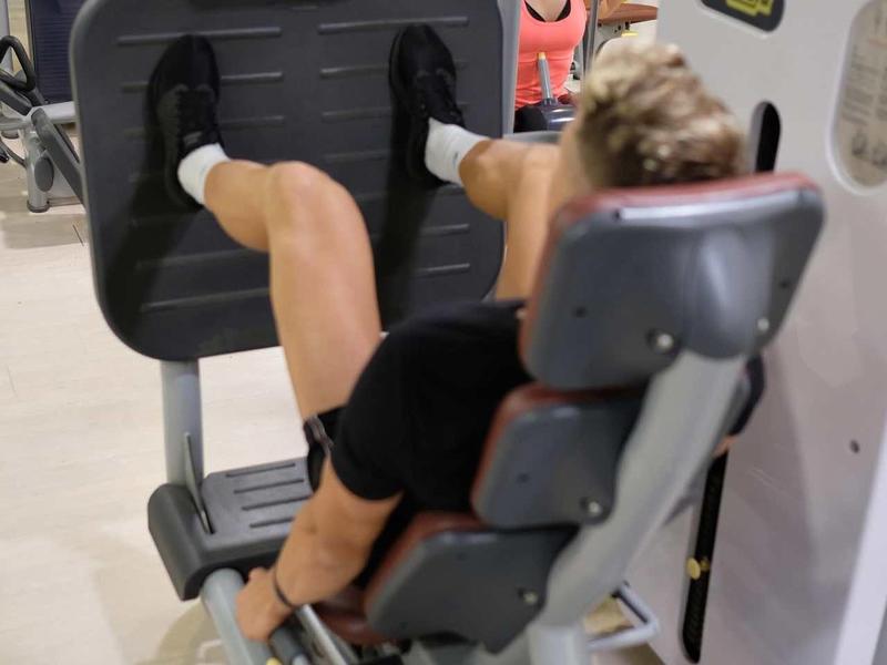 Woman exercising on leg press machine in a gym with mirrors and other equipment.