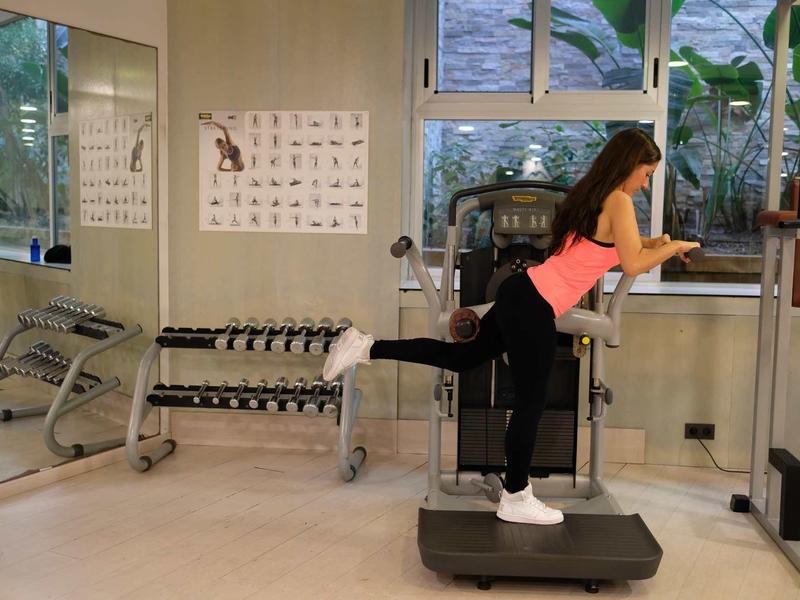 Woman exercising on a vibration platform in a bright gym with mirrored walls and dumbbells.