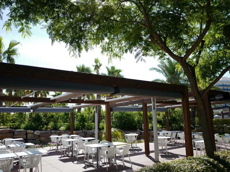 Outdoor seating area with white tables and chairs under leafy trees and pergolas.