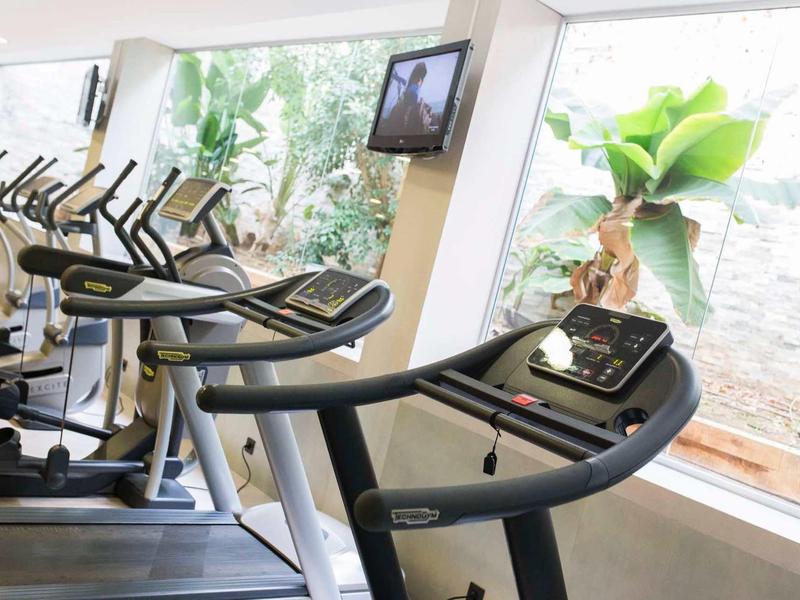 Indoor gym with treadmills and elliptical machines near large windows with plants outside.