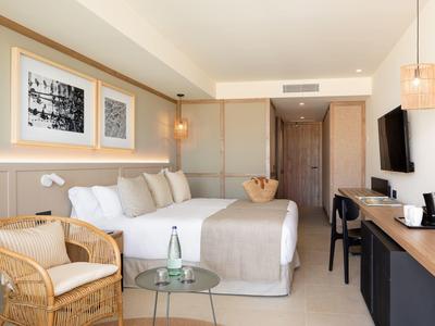 Camera d'hotel moderna con letto matrimoniale, sedia in vimini, tavolo in vetro e TV a muro.