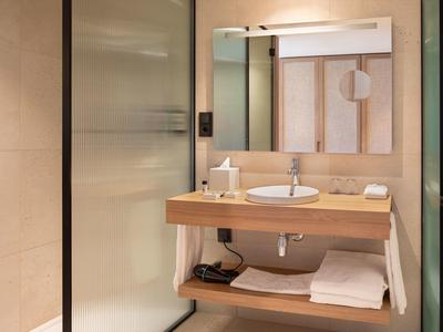 Bagno moderno in hotel con mobile in legno, lavandino bianco, grande specchio e doccia in vetro.