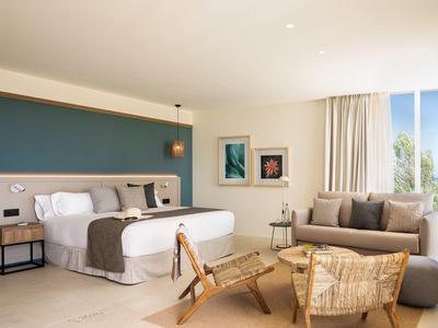 Camera d'hotel luminosa con letto, divano, sedie in rattan e grande finestra con vista sul verde.