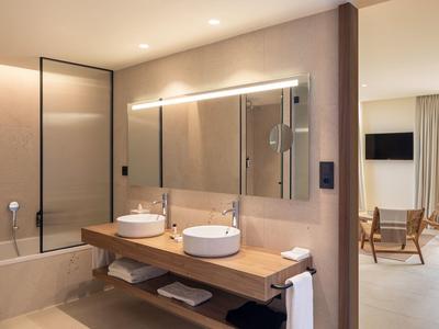 Bagno moderno in hotel con doppi lavandini, grande specchio, vasca da bagno e vista su un'area soggiorno accogliente.