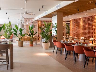 Ampio ristorante con tavoli in legno, sedie rosse, grandi piante e illuminazione calda.