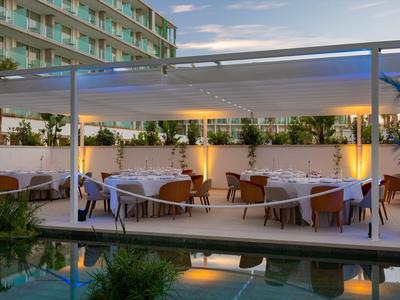 Area pranzo all'aperto con tavoli e sedie sotto una pergola vicino a una piscina al crepuscolo.