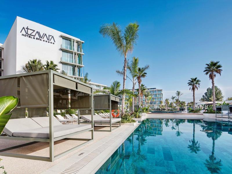 Hotel moderno con piscina all'aperto, lettini e palme sotto un cielo azzurro.