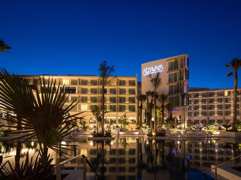 Hotel di lusso illuminato di notte con piscina e palme sotto un cielo blu intenso.