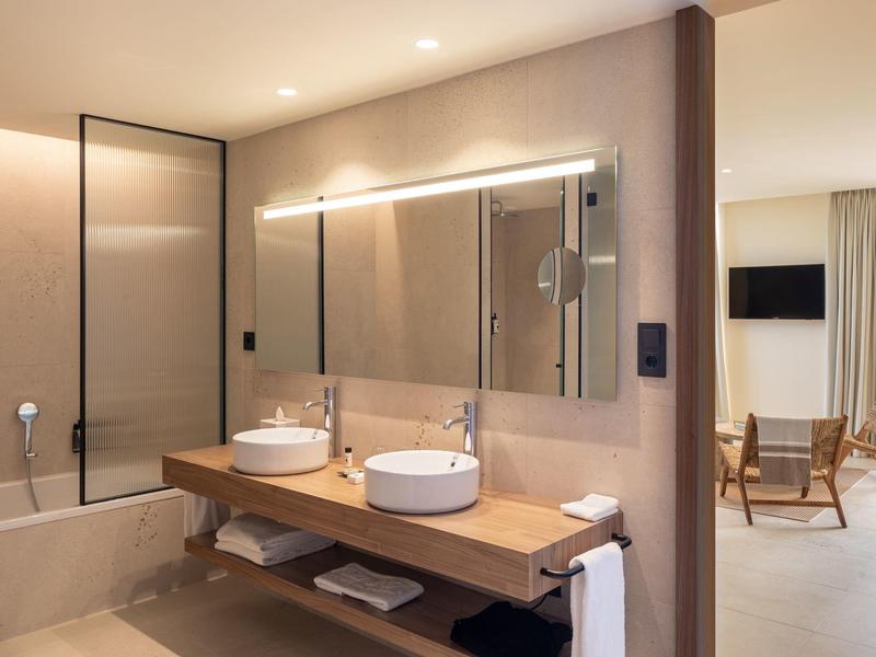 Bagno moderno in hotel con doppi lavandini, grande specchio, vasca da bagno e vista su un'area soggiorno accogliente.