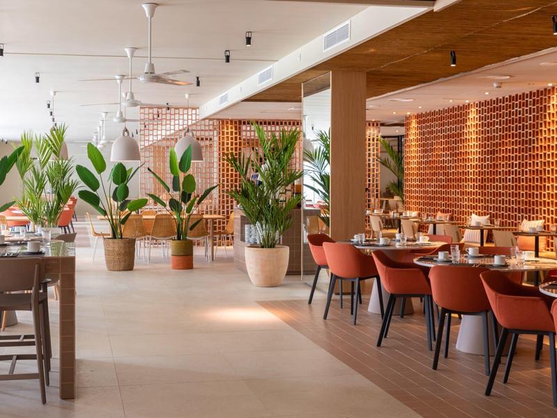 Ampio ristorante con tavoli in legno, sedie rosse, grandi piante e illuminazione calda.