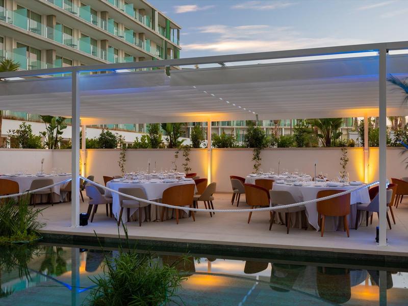 Area pranzo all'aperto con tavoli e sedie sotto una pergola vicino a una piscina al crepuscolo.