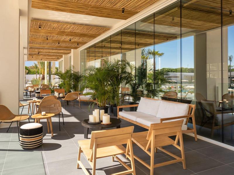 Terrazza moderna dell'hotel con mobili in legno, piante e pareti in vetro sotto un soffitto in legno.