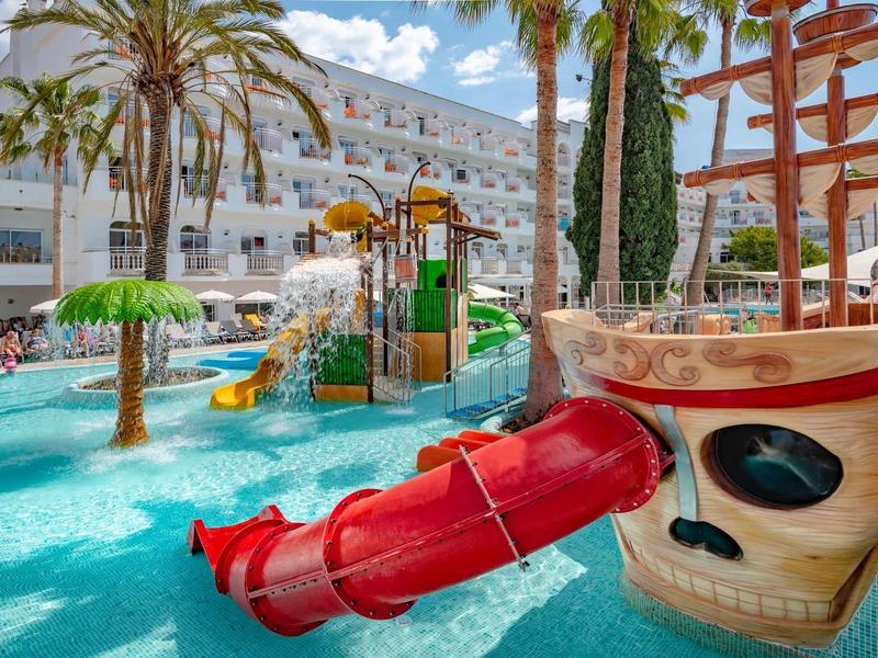 Colorida área de juegos acuáticos con toboganes, palmeras y un barco en la zona de piscina del hotel.