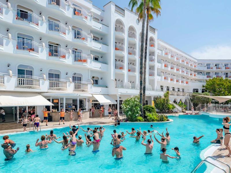 Hotel con gran piscina, muchos huéspedes practicando deportes acuáticos y palmeras bajo cielo azul.